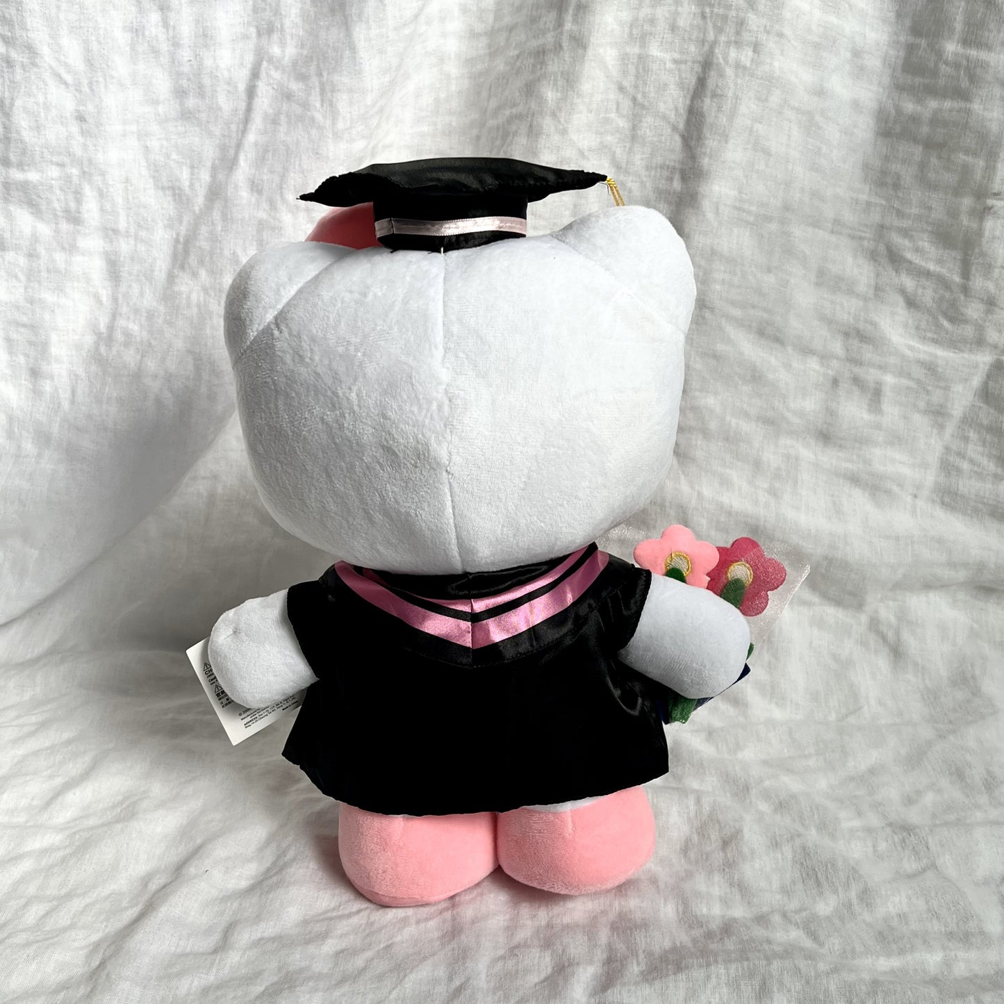 Back view of Hello Kitty graduation plush toy wearing a cap and gown, , against a grey background.