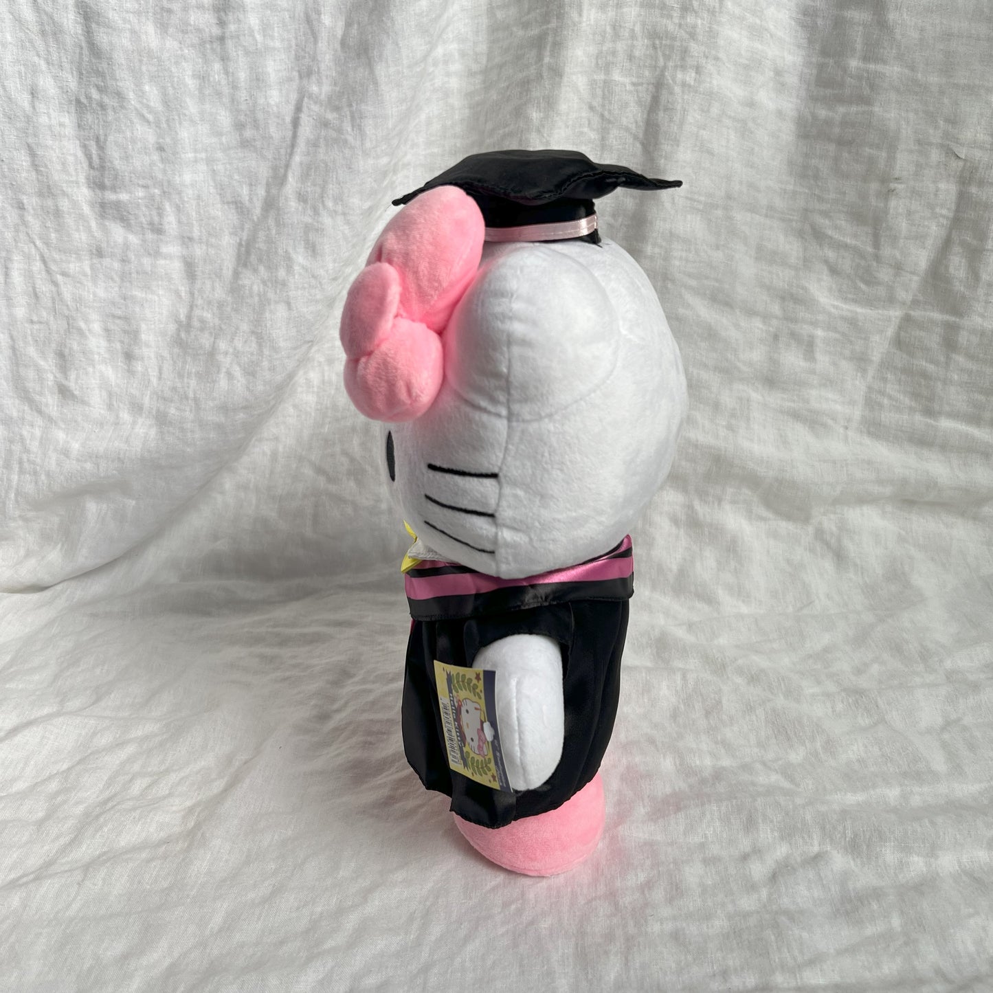 Side view of Hello Kitty graduation plush toy wearing a cap and gown, with licensing tag, against a grey background.