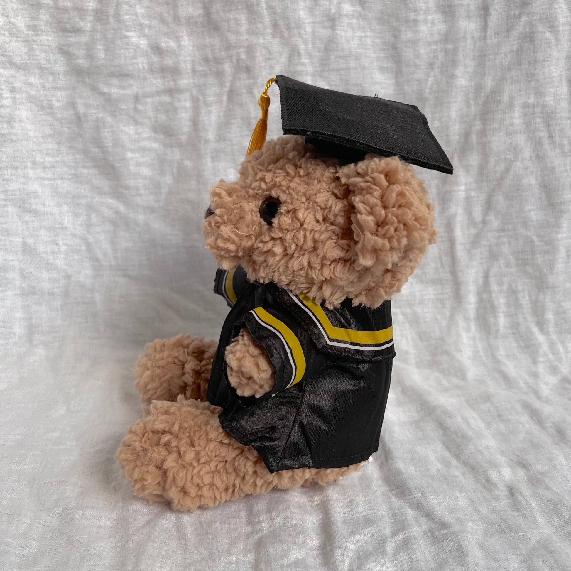 Front view of fluffy brown bear with graduation cap and gown containing yellow tassles and stripes