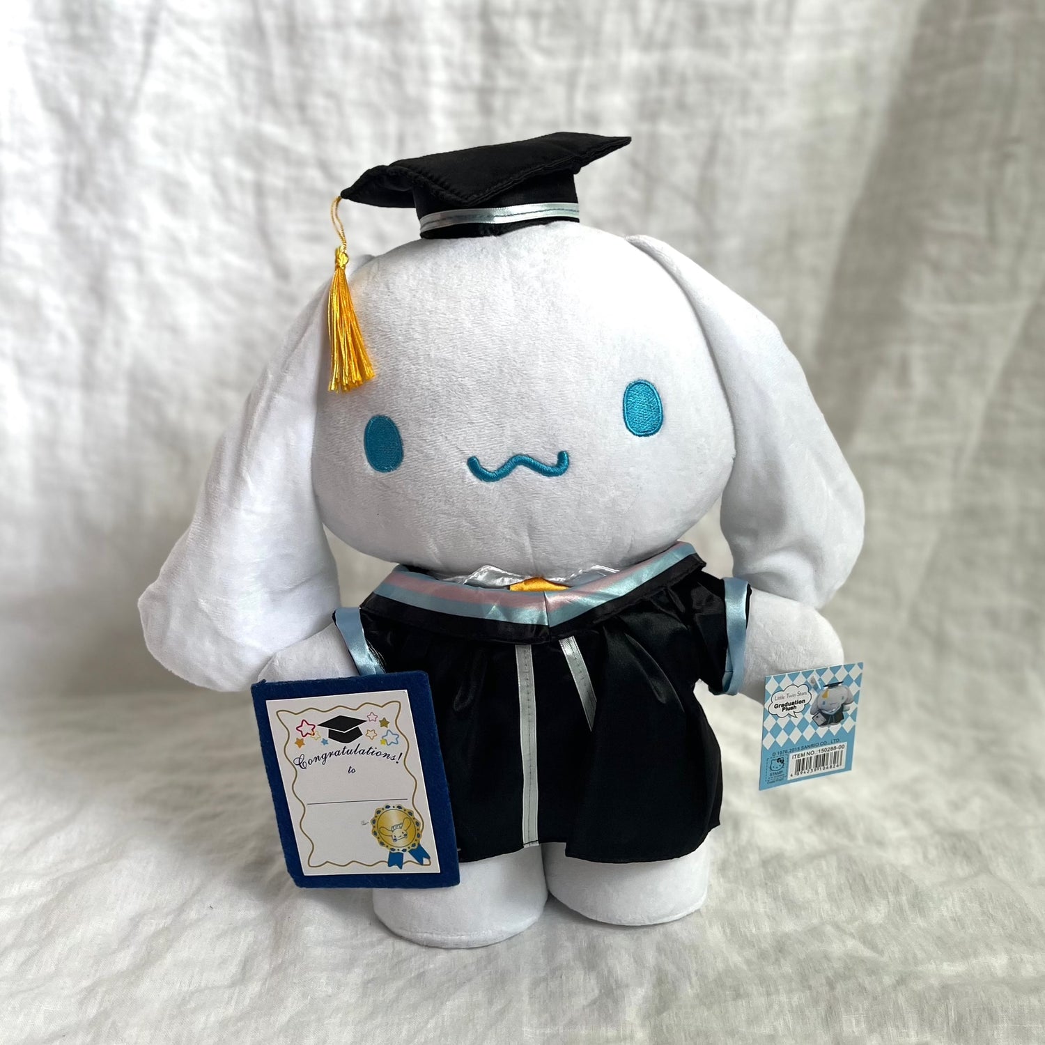 Front view of Cinnamoroll graduation gift featuring hat and gown with diploma in her hand. Golden tassles on the hat and light blue strips on the gown