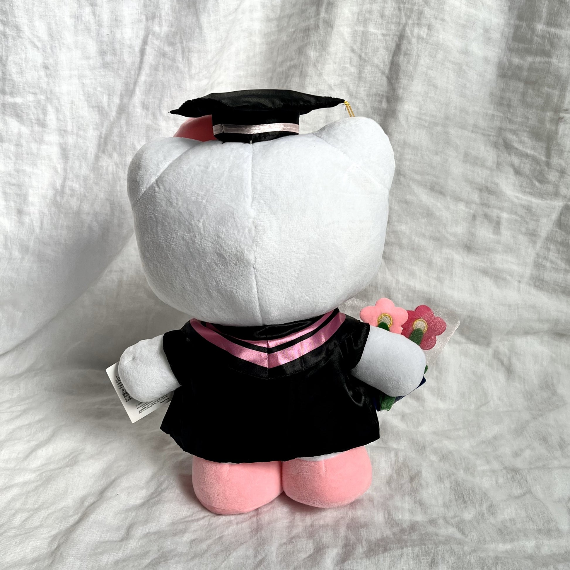 Back view of Hello Kitty graduation plush toy wearing a cap and gown, , against a grey background.