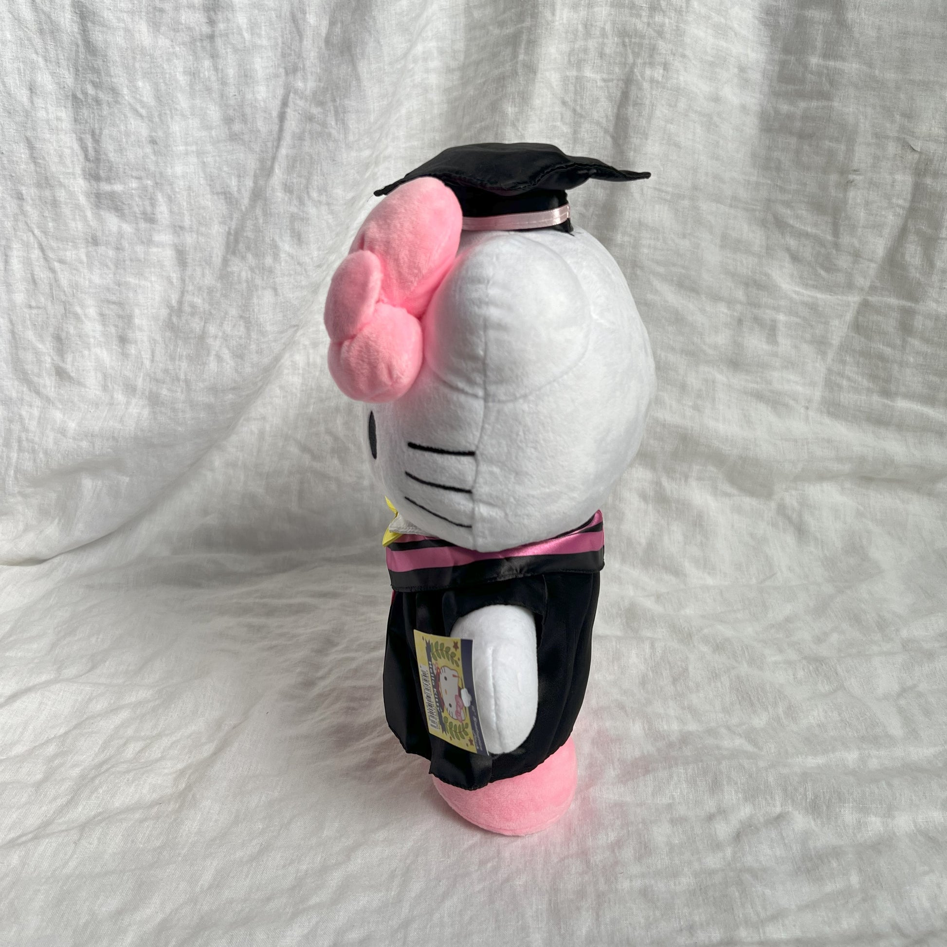 Side view of Hello Kitty graduation plush toy wearing a cap and gown, with licensing tag, against a grey background.